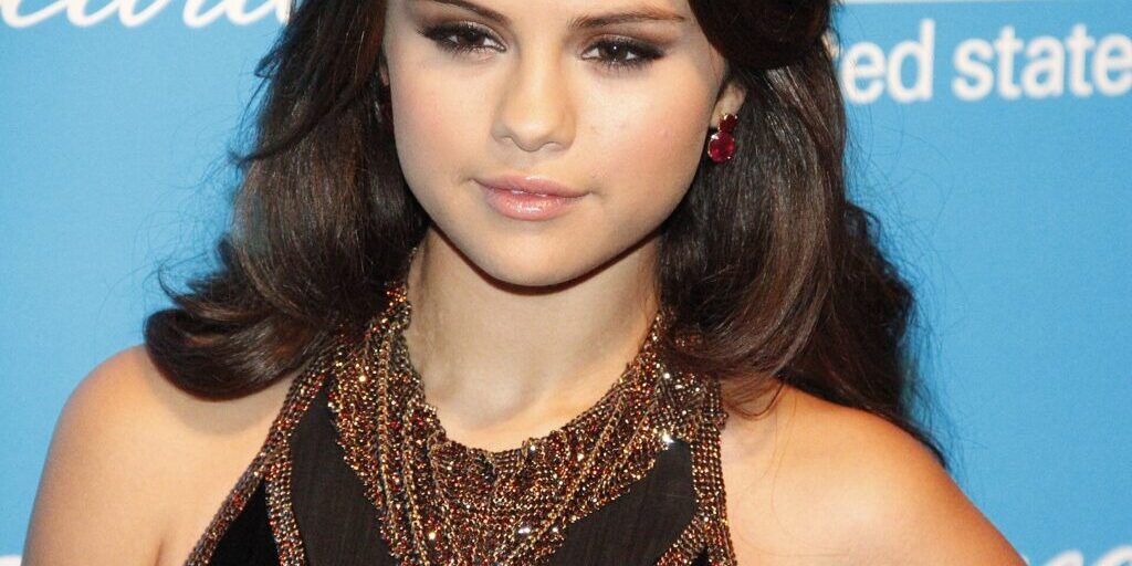 Selena Gomez in a black dress with gold embellishment, posing in front of a blue UNICEF backdrop.